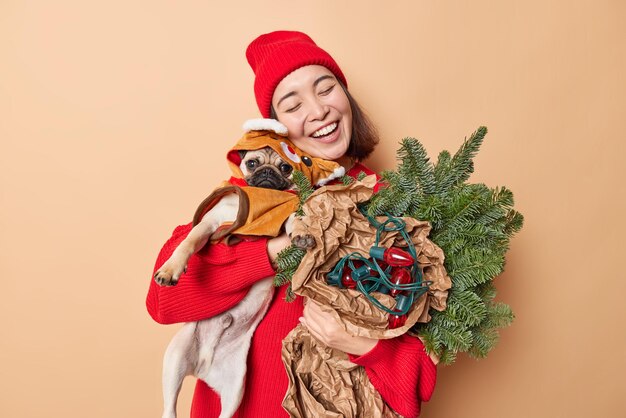 La donna felice e amichevole abbraccia il cane carlino con amore indossa il cappello rosso e il maglione porta attributi per la decorazione si sente felice di avere un'atmosfera festosa isolata su sfondo marrone. Concetto di vacanze invernali.