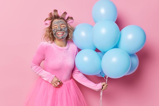 La donna felice sorride ampiamente subisce procedure di bellezza applica maschera di argilla e bigodini vuole avere una prospettiva perfetta indossa un vestito tiene un mazzo di palloncini blu si prepara per la festa Celebrazione