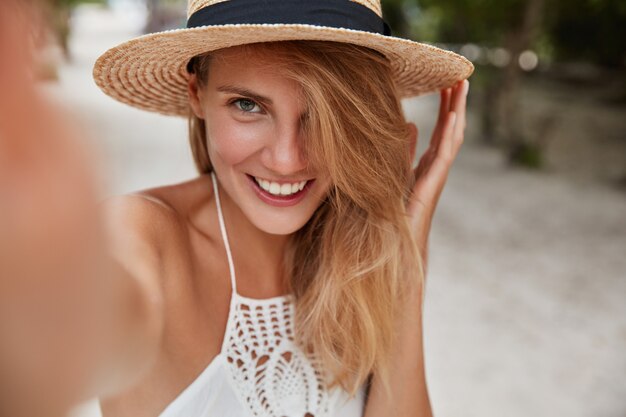 La donna gioiosa e spensierata indossa un cappello estivo di paglia alla moda sul dispositivo moderno e fa selfie, passeggia all'aperto durante il soleggiato clima estivo. Persone, stile di vita e concetto di riposo.