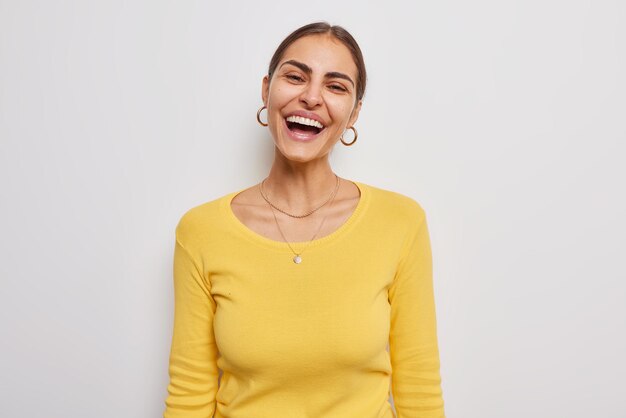 La donna gioiosa felice sorride ampiamente mostra i denti bianchi che indossa orecchini e il maglione giallo casual si sente in pose positive al chiuso su sfondo bianco si sente una buona notizia. Concetto di persone ed emozioni.