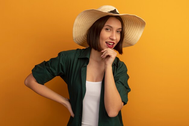 La donna graziosa allegra con il cappello della spiaggia mette la mano sul mento e guarda la parte anteriore isolata sulla parete arancione