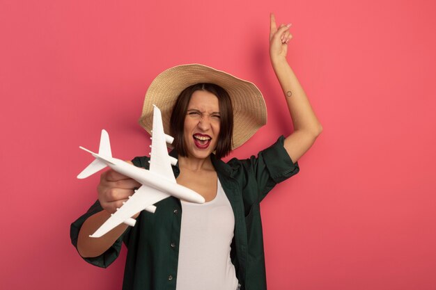 La donna graziosa allegra con il cappello della spiaggia tiene l'aereo del modello e indica in alto isolato sulla parete rosa