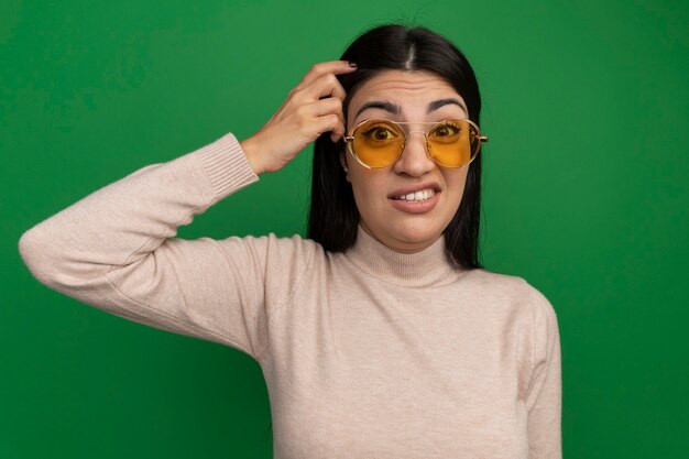La donna graziosa del brunette confusa in occhiali da sole mette il dito sulla testa isolata sulla parete verde