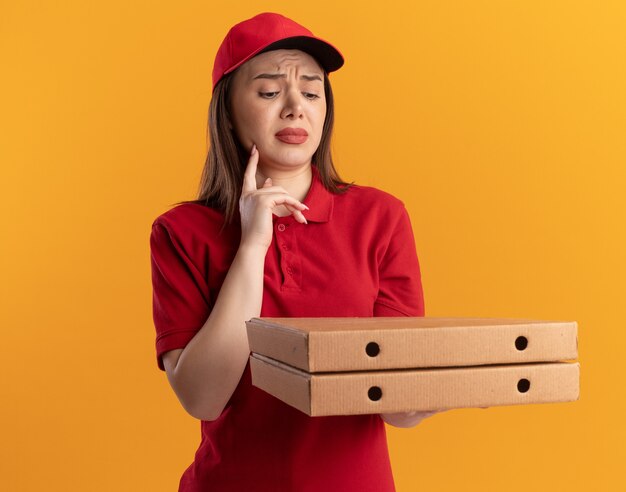 La donna graziosa di consegna delusa in uniforme mette il dito sul mento che tiene e guardando le scatole per pizza sull'arancia