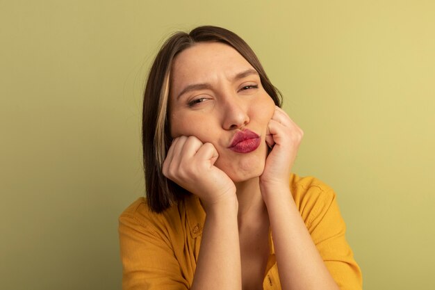 La donna graziosa offesa mette i pugni sulle guance isolate sulla parete verde oliva