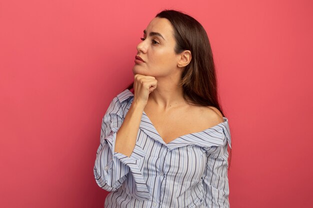 La donna graziosa sicura tiene il mento che esamina il lato isolato sulla parete rosa