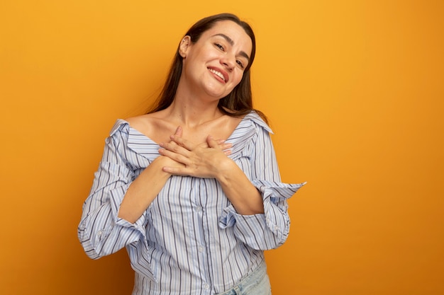 La donna graziosa sorridente mette le mani sul petto isolato sulla parete arancione