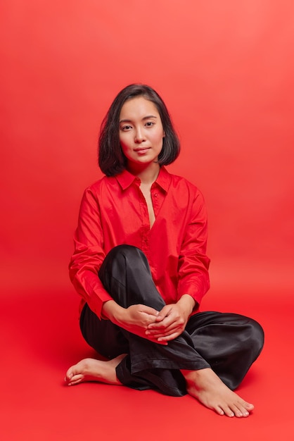 la donna guarda direttamente la telecamera indossa una camicia rossa e pantaloni neri posa a piedi nudi al coperto. Il modello femminile grazioso riposa le pose dell'interno allo studio che va a praticare lo yoga