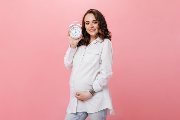 La donna incinta abbastanza giovane in camicia bianca sorride ampiamente e posa sull'isolato. Ragazza bruna tiene sveglia su sfondo rosa.
