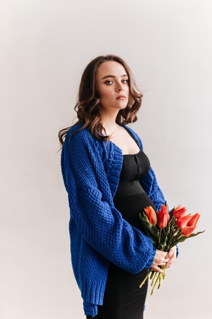 La donna incinta castana riccia in cardigan blu tiene il mazzo dei tulipani. Attraente signora in abito nero pone su sfondo isolato.