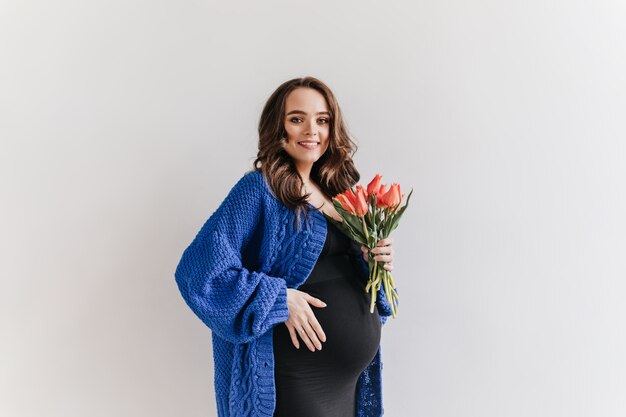 La donna incinta castana riccia in vestito nero e cardigan blu tiene i tulipani. Bella signora sorride su sfondo bianco.