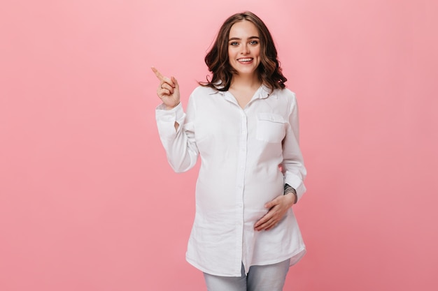 La donna incinta di buon umore tocca la pancia e il sorriso. Ragazza bruna in camicia bianca indica il luogo per il testo su sfondo rosa isolato.