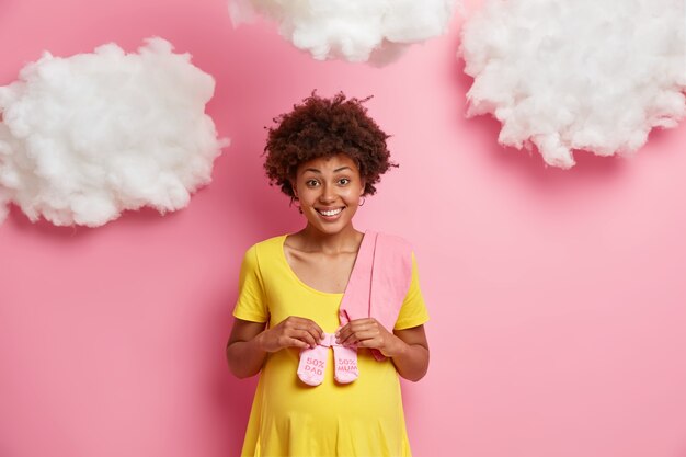 La donna incinta felice con i capelli afro tiene i calzini del bambino sopra la pancia si prepara a partorire