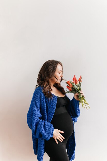 La donna incinta felice riccia in cardigan blu tiene i fiori. Affascinante ragazza bruna in abito nero odora il bouquet di tulipani su sfondo bianco.