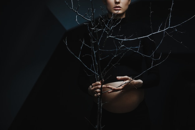 La donna incinta in vestiti neri si leva in piedi sotto la parete grigia e tiene un ramo di albero