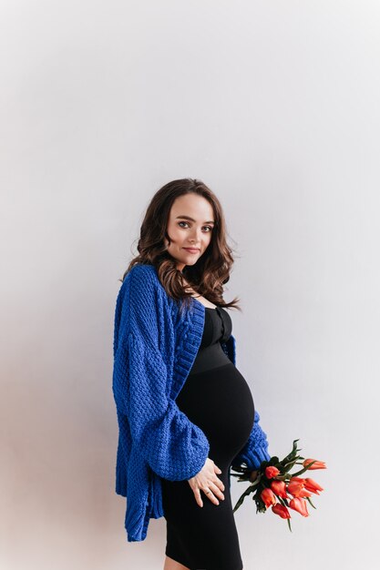 La donna incinta riccia castana in cardigan blu tiene il mazzo dei fiori. Affascinante signora in abito nero posa con tulipani su sfondo isolato.