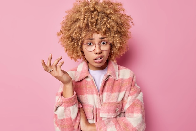 La donna indignata con i capelli ricci alza la mano e scrolla le spalle le borse delle labbra indossa una giacca a scacchi ha un'espressione esitante isolata su sfondo rosa Quindi cosa dovrei fare in questa situazione