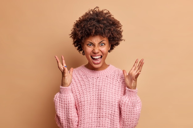 La donna indignata grida gesti con rabbia con le mani alzate esprime irritazione e urla ad alta voce indossa un maglione lavorato a maglia isolato sopra il muro marrone