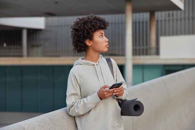 la donna indossa la felpa tiene i tipi di telefono cellulare i messaggi di testo trasportano il karemat arrotolato focalizzato lontano ha un allenamento di yoga ogni giorno