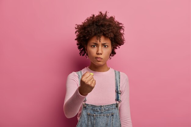 La donna infastidita insoddisfatta con i capelli afro sorride e stringe i pugni, guarda con rabbia qualcuno, promette di vendicarsi o punire per il cattivo comportamento, ha un'espressione infastidita, posa su un muro roseo