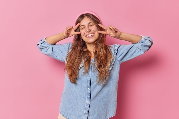 la donna invia vibrazioni positive fa un gesto di pace sugli occhi sorride a trentadue denti vestita con una camicia di jeans isolata sul rosa mostra il segno della vittoria ha un umore allegro. Linguaggio del corpo.