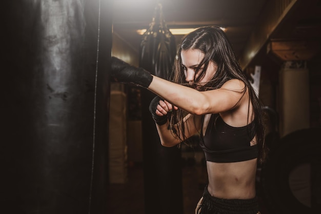 La donna magra e concentrata ha un allenamento di boxe con un sacco da boxe in una palestra oscura.