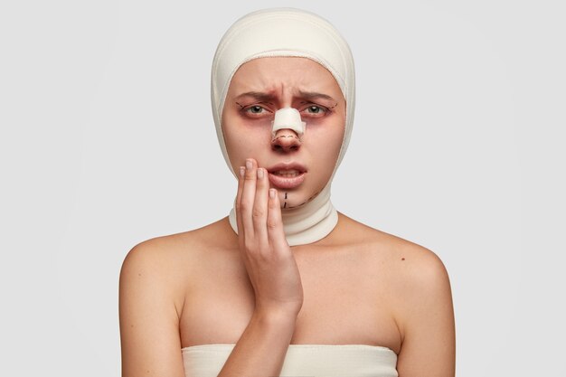La donna malata disperata tiene la mano sulla guancia, soffre di forti dolori dopo l'innesto di pelle, ha bendaggio o cerotto sul naso