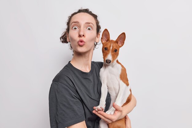 la donna mantiene le labbra arrotondate fissa con shock trattiene il cane basenji scatta una foto di se stessa e l'animale domestico fa una smorfia divertente isolata su bianco