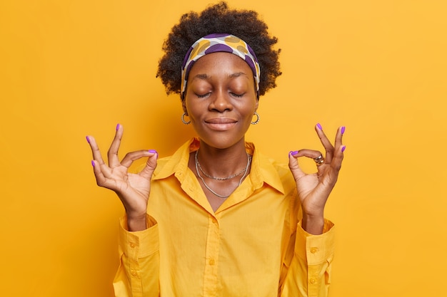 la donna medita con gli occhi chiusi pratica lo yoga tiene le mani in un gesto ok indossa una camicia casual posa su un giallo vivido