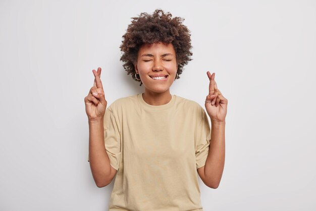 la donna morde le labbra tiene le dita incrociate crede nella buona fortuna indossa una maglietta beige casual isolata su bianco. Possano i miei sogni diventare realtà