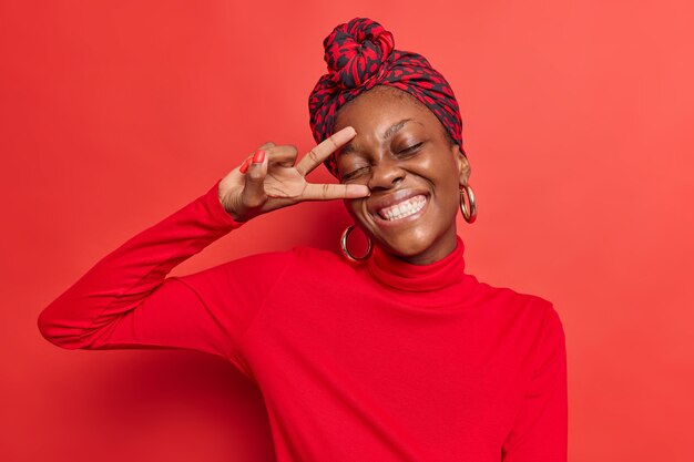 la donna mostra il segno v vicino al viso gesto di pace tiene gli occhi chiusi sorrisi ampiamente vestita posa casualmente su studio rosso brillante