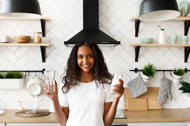 La donna mulatta sorrisa con i capelli sciolti tiene in mano un bicchiere vuoto e un bicchiere di latte vicino allo scrittorio della cucina sulla moderna cucina bianca vestita di t-shirt bianca