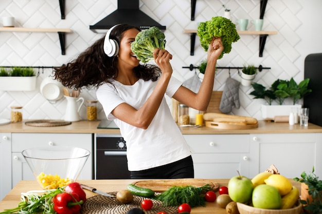 La donna mulatta sorrisa in grandi cuffie senza fili sta ballando con foglie di insalata e broccoli sulla cucina moderna vicino al tavolo pieno di frutta e verdura
