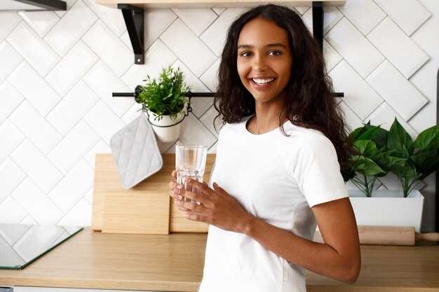 La donna mulatta sorrisa sta tenendo il bicchiere d'acqua vicino alla scrivania della cucina moderna bianca vestita con una maglietta bianca