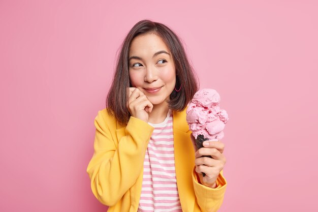 La donna orientale sognante guarda piacevolmente lontano con un'espressione premurosa tiene un grande gelato gustoso pensa a qualcosa che tiene la mano sul viso vestito con abiti eleganti isolati sul muro rosa