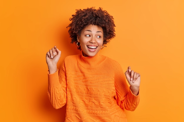 La donna ottimista balla con le braccia alzate muove sorrisi spensierati ampiamente vestiti in un maglione casual isolato su un muro arancione vivido celebra il successo e la vittoria gode di una bella giornata