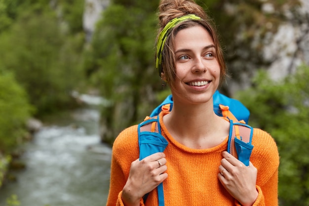 La donna pensosa e felice viaggia in un luogo maestoso, guarda felicemente lontano, indossa un maglione arancione casual, porta lo zaino