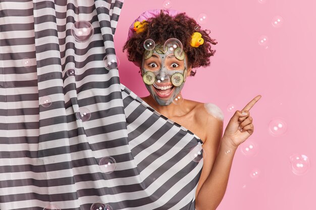 La donna positiva con i capelli ricci applica una maschera all'argilla per il ringiovanimento della pelle contro le bolle di sapone rosa della parete intorno. Guarda questo prodotto per l'igiene