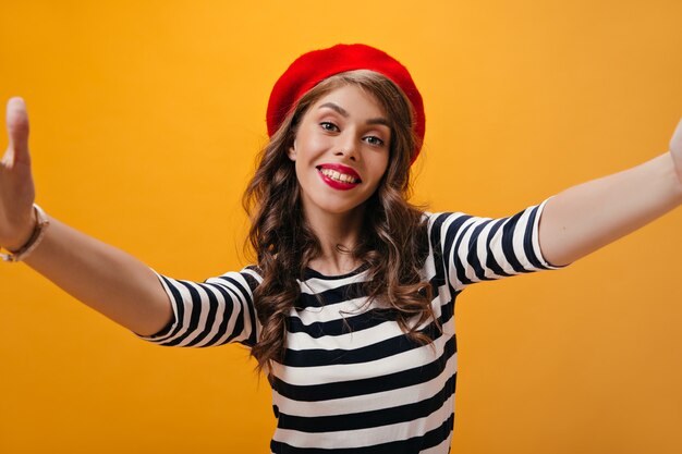 La donna positiva in camicia a righe e berretto luminoso fa selfie. Cool ragazza giovane con acconciatura moderna in cappello rosso scatta foto su sfondo isolato.
