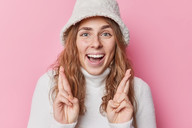 La donna positiva speranzosa tiene le dita incrociate e fa desiderare o anticipa qualcosa sorride attende con gioia i desideri di un risultato positivo per la vittoria vestita con abiti invernali isolati su sfondo rosa