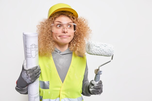 la donna preme le labbra focalizzate sulla distanza indossa l'uniforme del casco protettivo e gli occhiali di sicurezza essendo un decoratore professionista tiene le pose del modello del rullo al coperto. Servizio di riparazione a domicilio