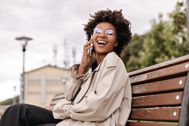 La donna riccia castana eccitata felice in trench beige e occhiali da vista ride, parla al telefono e si siede sulla panca di legno fuori