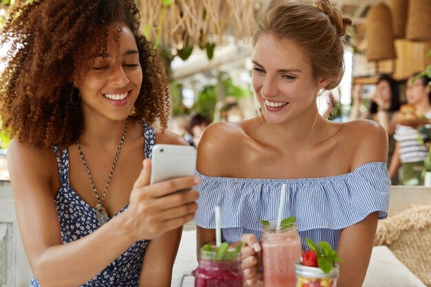 La donna riccia dalla pelle scura con espressione positiva mostra le foto alla sua migliore amica su smart phone, beve frullato. Coppia lesbica ricreare nel ristorante con gadget moderni. Concetto di amicizia.