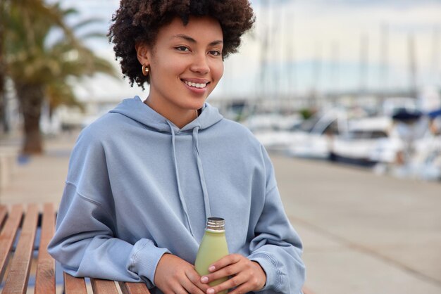 la donna riposa dopo l'allenamento sportivo nel porto si siede su una panca di legno ammira viste fantastiche beve acqua fresca indossa una felpa si sente rilassata osserva la baia della città con gli yacht