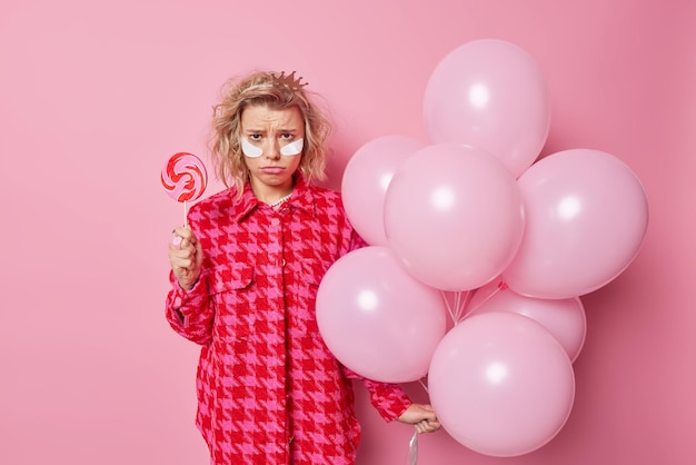 La donna sconvolta frustrata ha cattivo umore dopo la festa tiene deliziose caramelle rotonde sul bastone e un mucchio di palloncini gonfiati indossa toppe di bellezza vestite con abiti alla moda isolati sopra il muro rosa