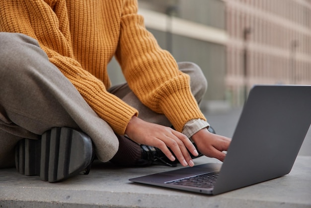 La donna senza volto si siede sui tipi di gambe incrociate sul moderno computer portatile indossa un caldo maglione lavorato a maglia installa l'applicazione dal sito web di navigazione in rete connesso al wifi Freelace e il concetto di lavoro a distanza