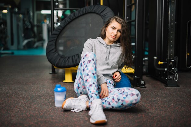 La donna seria atletica che si siede vicino imbottiglia la palestra