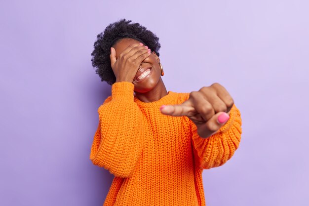 La donna si diverte ride felicemente contro gli occhi con la mano punta il dito indice in telecamera vede qualcosa di divertente indossa un maglione lavorato a maglia arancione isolato su viola