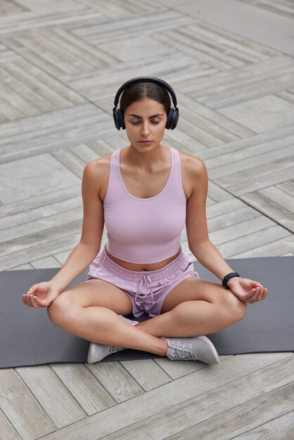 la donna si siede nella posizione del loto conduce uno stile di vita atletico si rilassa dopo una giornata intensa ascolta musica per rilassarsi in cuffie wireless indossa pantaloncini e scarpe da ginnastica respira profondamente