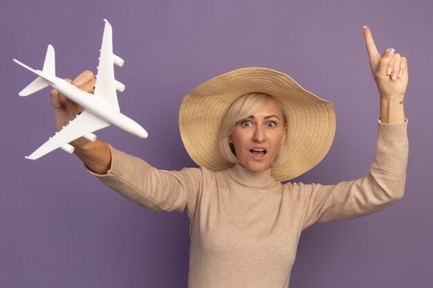 La donna slava abbastanza bionda sorpresa con il cappello della spiaggia tiene l'aereo del modello e indica la porpora